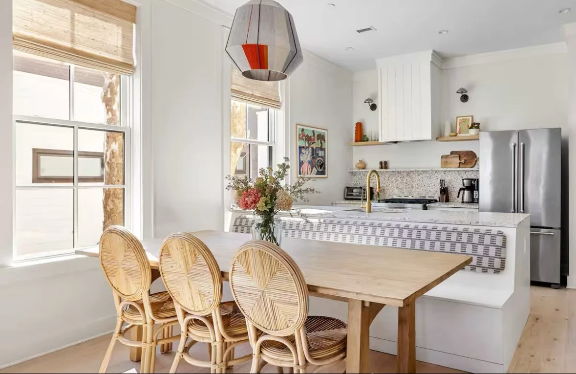 Bright, modern kitchen with white cabinets, granite countertops, stainless steel refrigerator, and a dining table with four rattan chairs and a floral centerpiece.