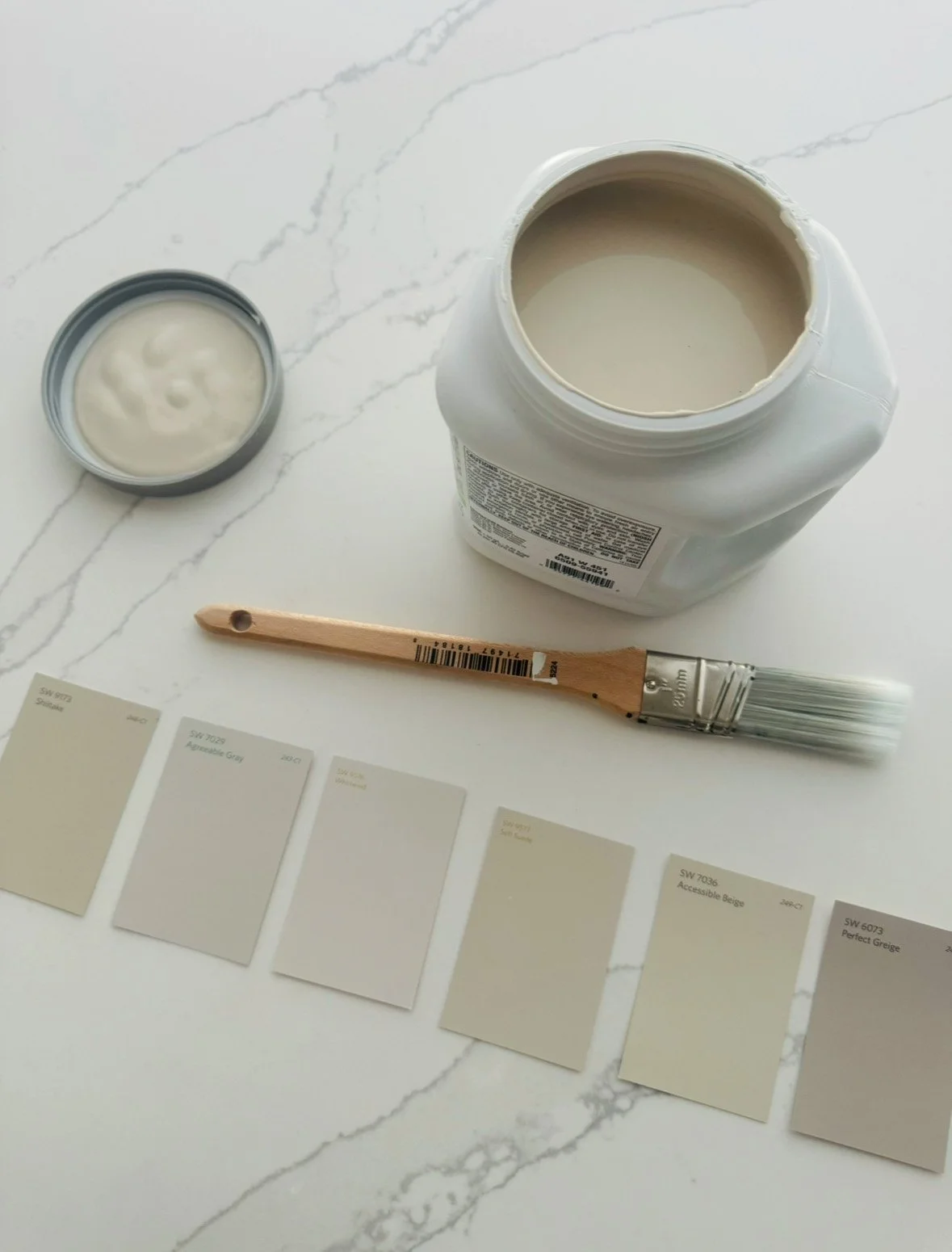 A paint color matching station with a container of light beige paint, a paintbrush, a can of beige paint sample, and color swatches with various shades of beige and gray on a white marble surface.