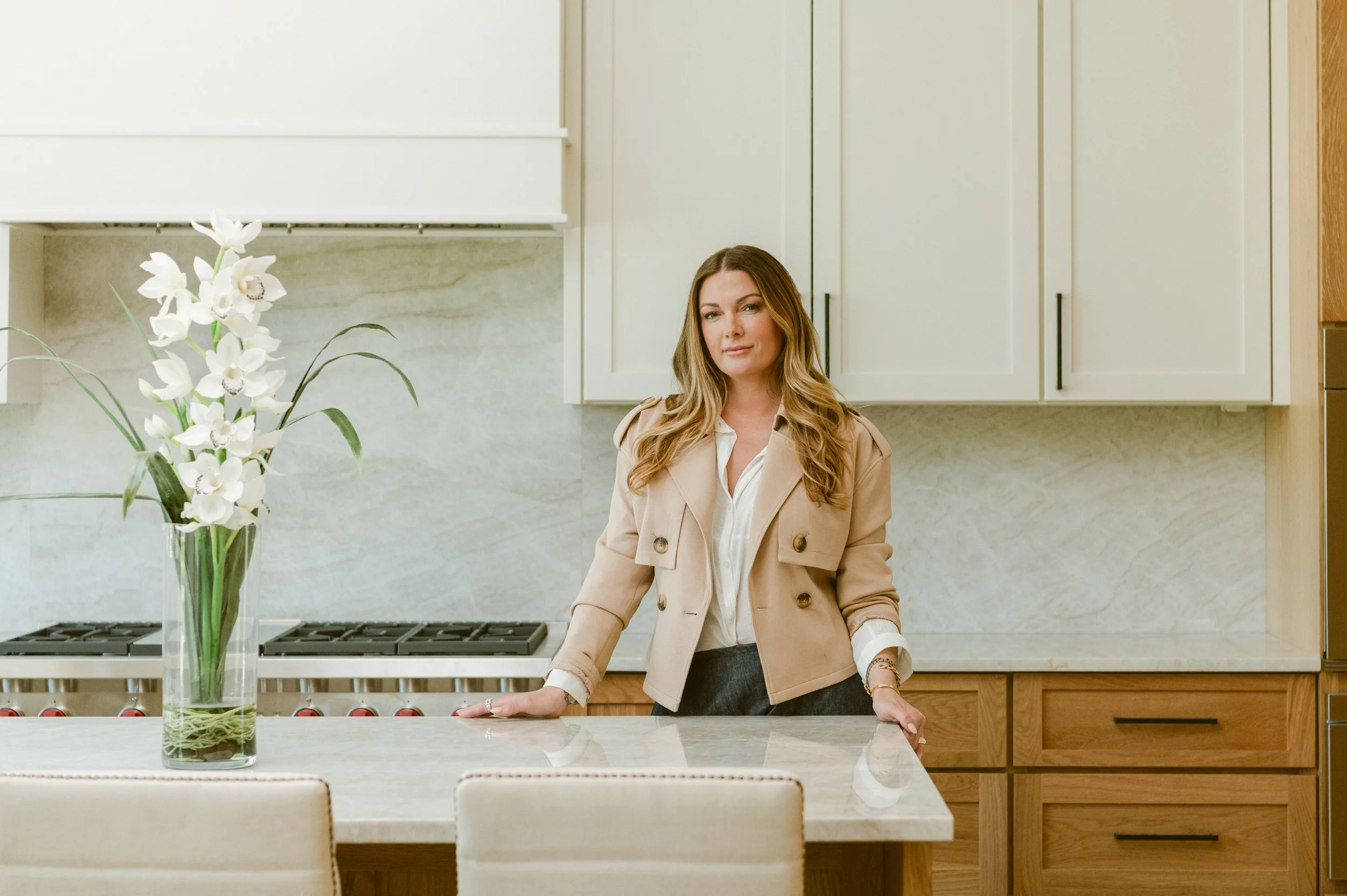 Lisa with long wavy hair wearing a beige jacket and white shirt standing in a modern kitchen with white cabinets and a marble countertop, near a large glass vase with white orchids.