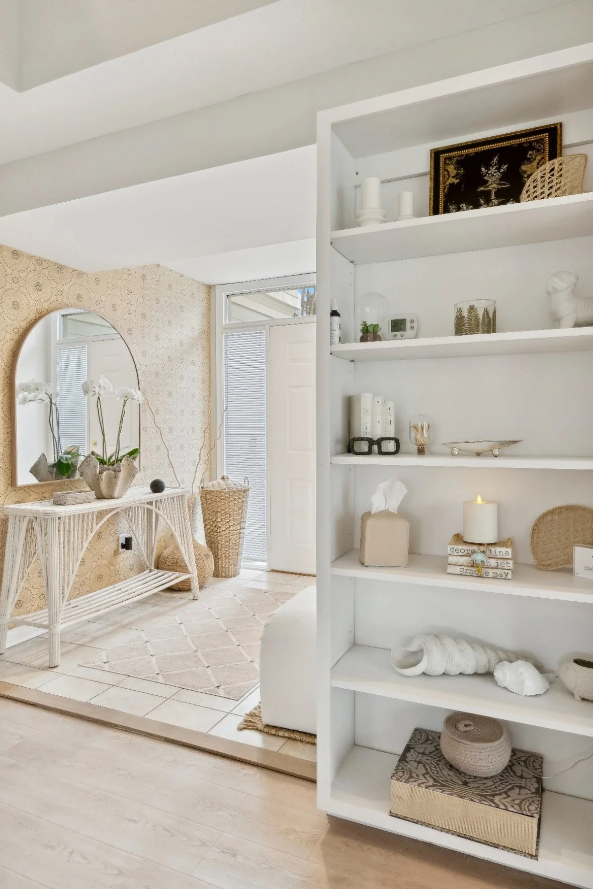 A bright, airy entryway with a white shelving unit filled with neutral-colored decorative items, a mirror on a textured beige wall, and a white console table with orchids and wicker baskets.