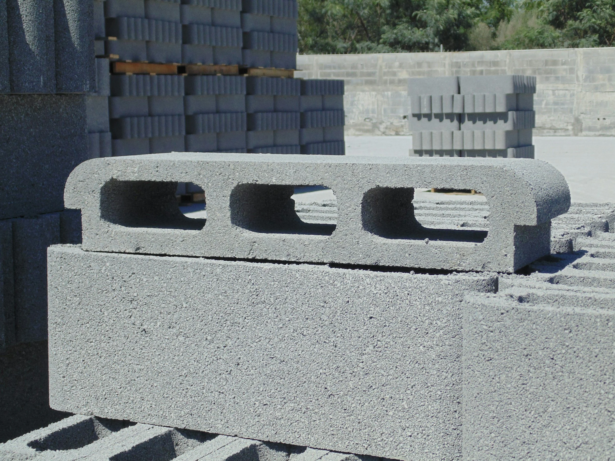 A stack of gray concrete cinder blocks outdoors with other stacks of similar blocks in the background, set against trees and a concrete wall.