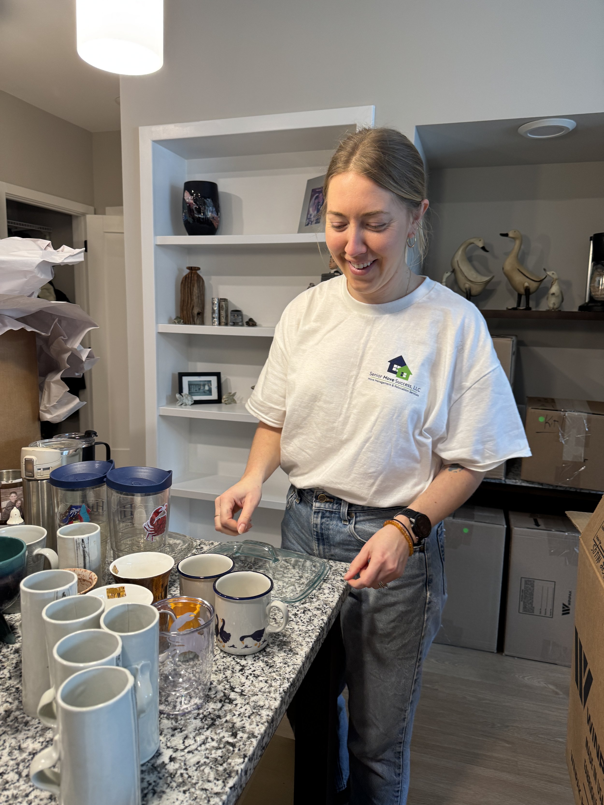 Bright living room with beige curtains, white walls, a gray and white sofa with textured pillows, a white armchair, a round marble coffee table with vases and books, a wooden sideboard, a large potted plant, wall art, and natural light from a floor-to-ceiling window showing trees outside.