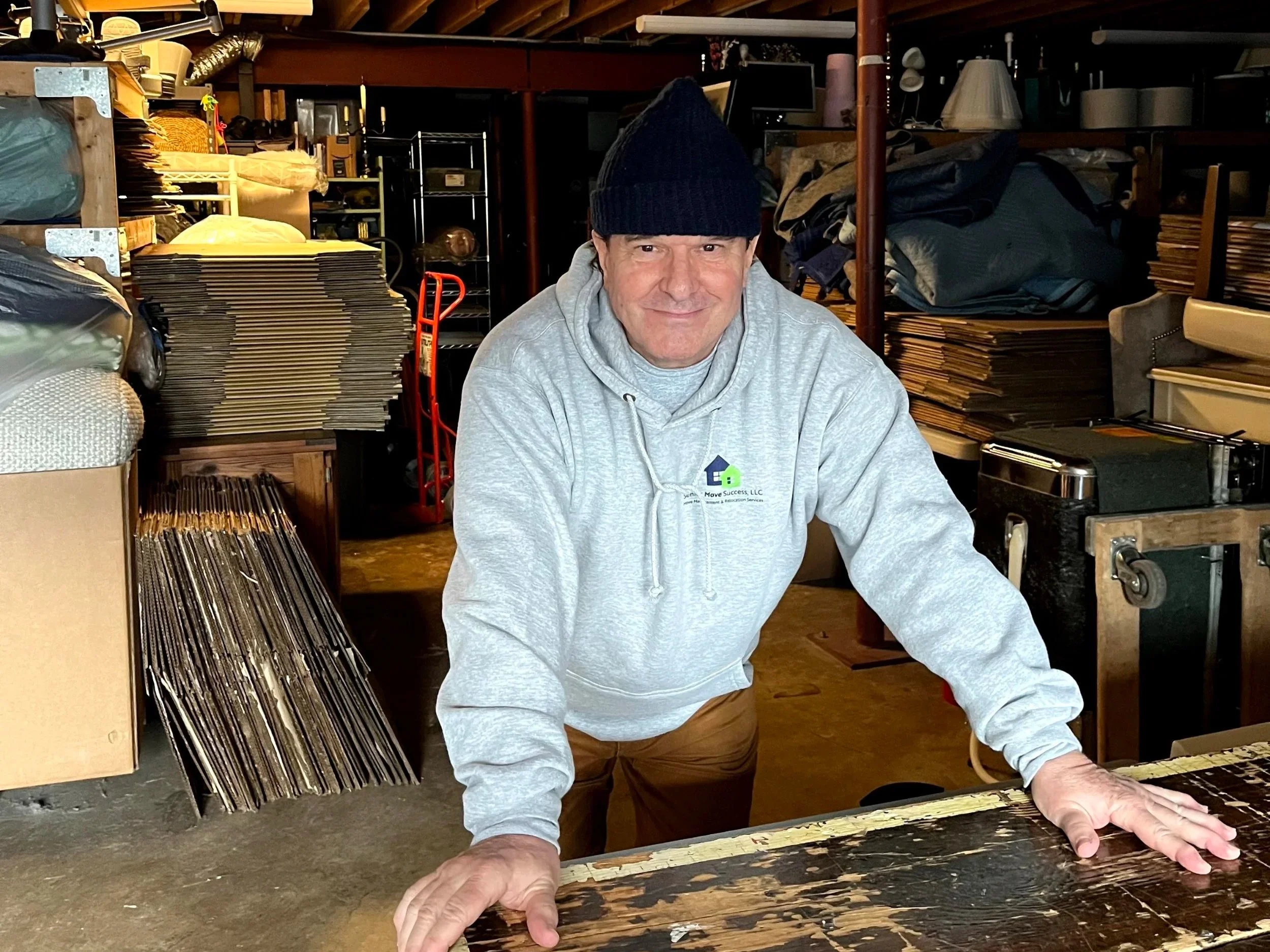 A collection of cardboard boxes, potted plants, and furniture wrapped for moving inside a bright room with large windows.