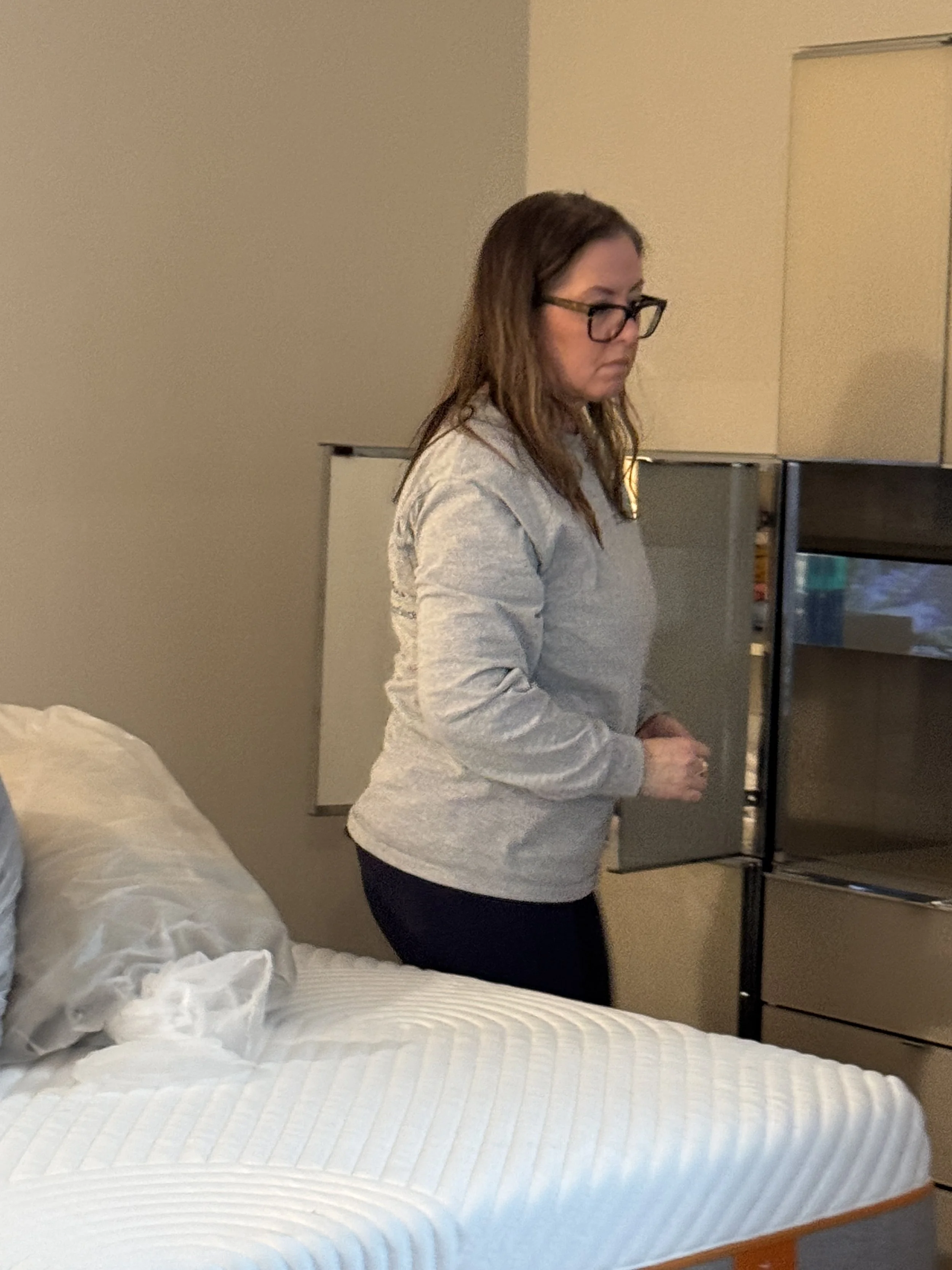 A woman with glasses and brown hair standing next to a bed in a hotel room, looking at a refrigerator.