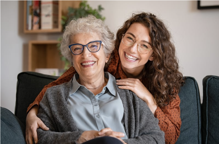 Two women, an elderly woman with gray hair and glasses and a younger woman with curly brown hair, smiling and sitting close together on a couch in a living room.