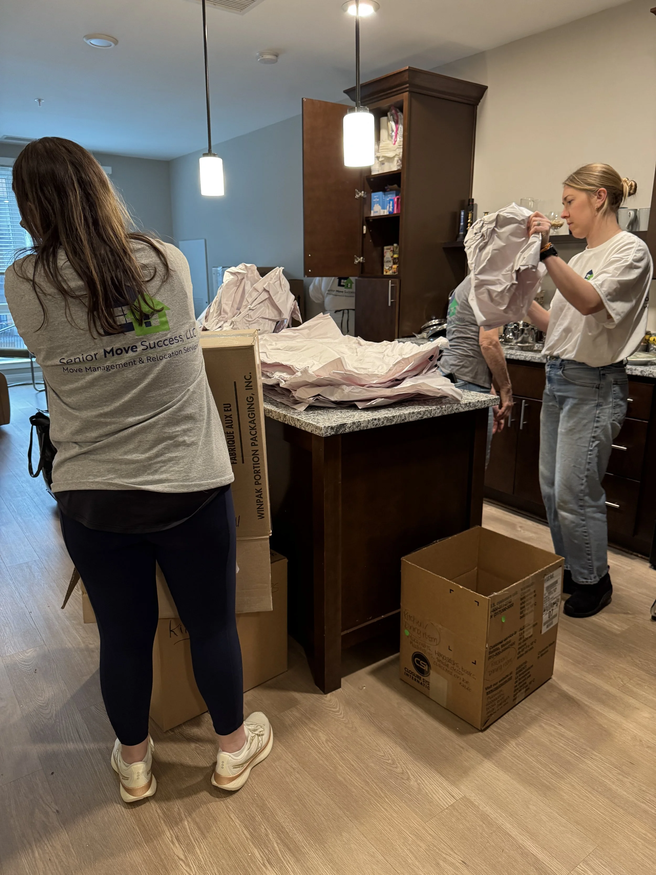 Three women packing or unpacking boxes in a kitchen with brown cabinets and wooden floors.