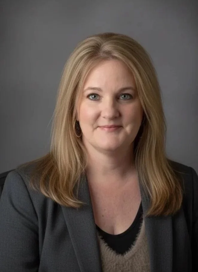 A woman with shoulder-length blonde hair wearing a gray blazer and black top, smiling against a gray background.