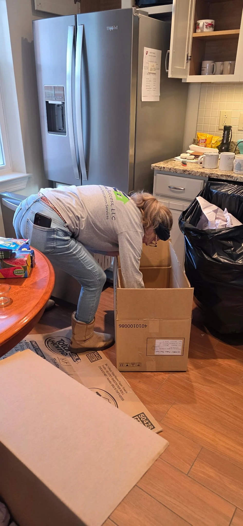 A woman is bending into a cardboard box in a kitchen, wearing jeans, a gray t-shirt, tan boots, and a blindfold.