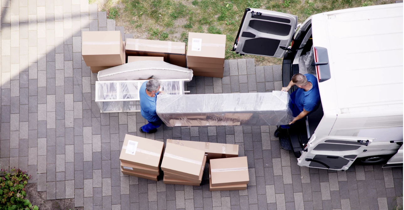 Two movers loading a large wrapped piece of furniture into a white delivery van with open doors, surrounded by boxes on a brick driveway.