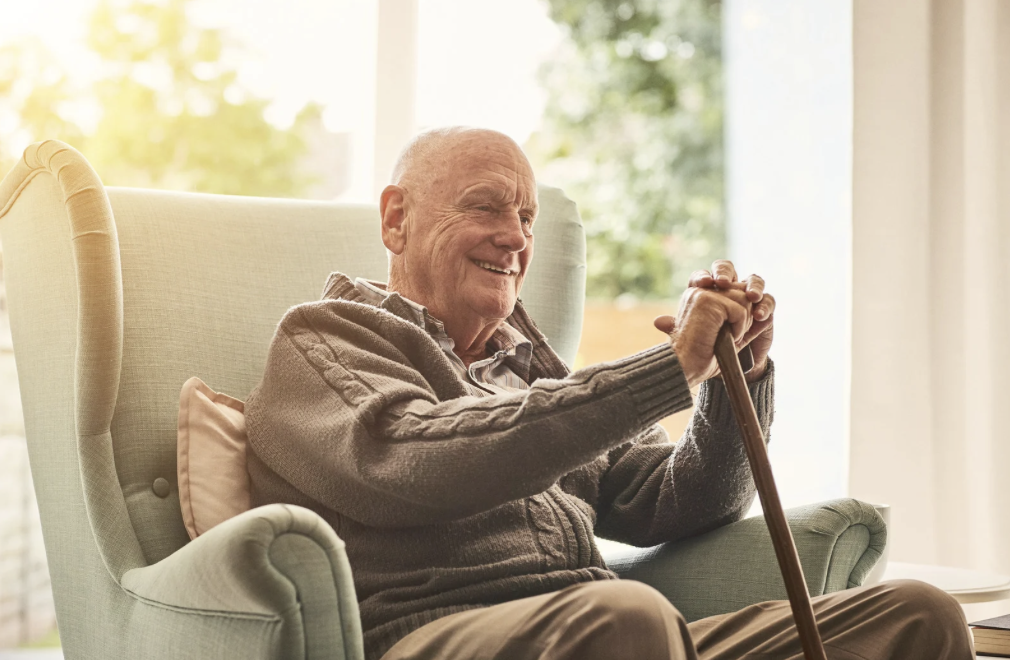 An elderly man sitting in a comfortable armchair, smiling, holding a wooden cane with both hands, with large windows and greenery in the background.