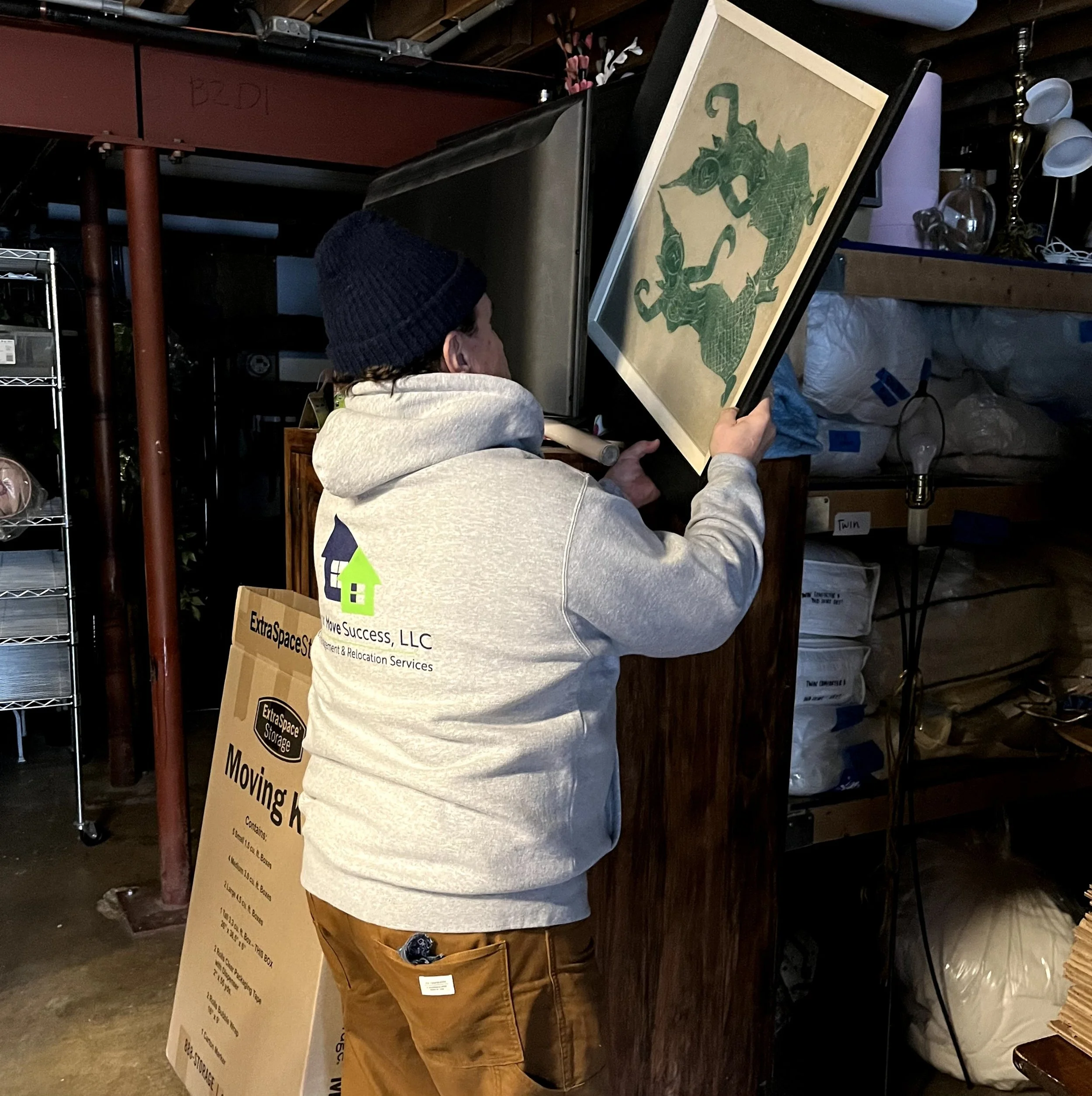 A person wearing a gray hoodie and a dark knit hat, facing away, holding a framed piece of art with a dragon design, in a storage or moving space.