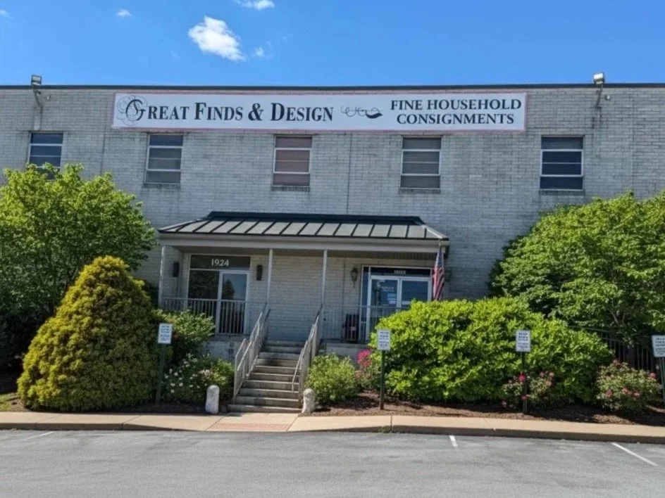 Exterior of a white brick building with a sign reading 'Great Finds & Design, Fine Household Consignments'. The building has four windows, a small staircase leading to the entrance, and an American flag displayed to the right of the door. There are green bushes and trees in front of the building, and a parking lot in the foreground.