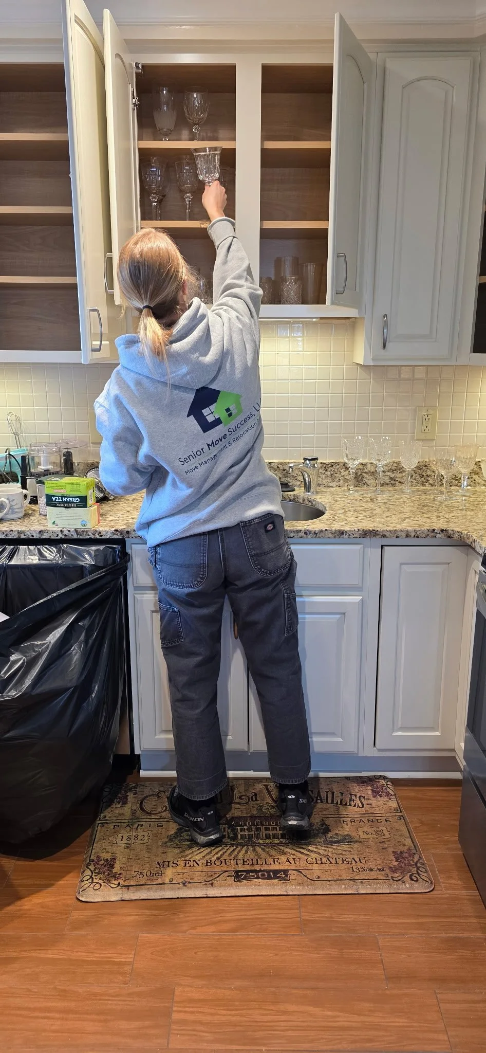 A person wearing a gray hoodie and jeans is reaching into a top kitchen cabinet that contains glassware. The person is standing on a decorative rug in a kitchen with beige cabinets, a granite countertop, and a tiled backsplash. There are glass cups o
