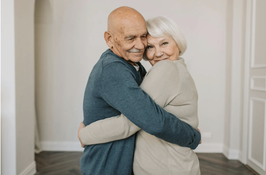 An elderly couple hugging and smiling in a cozy indoor setting.