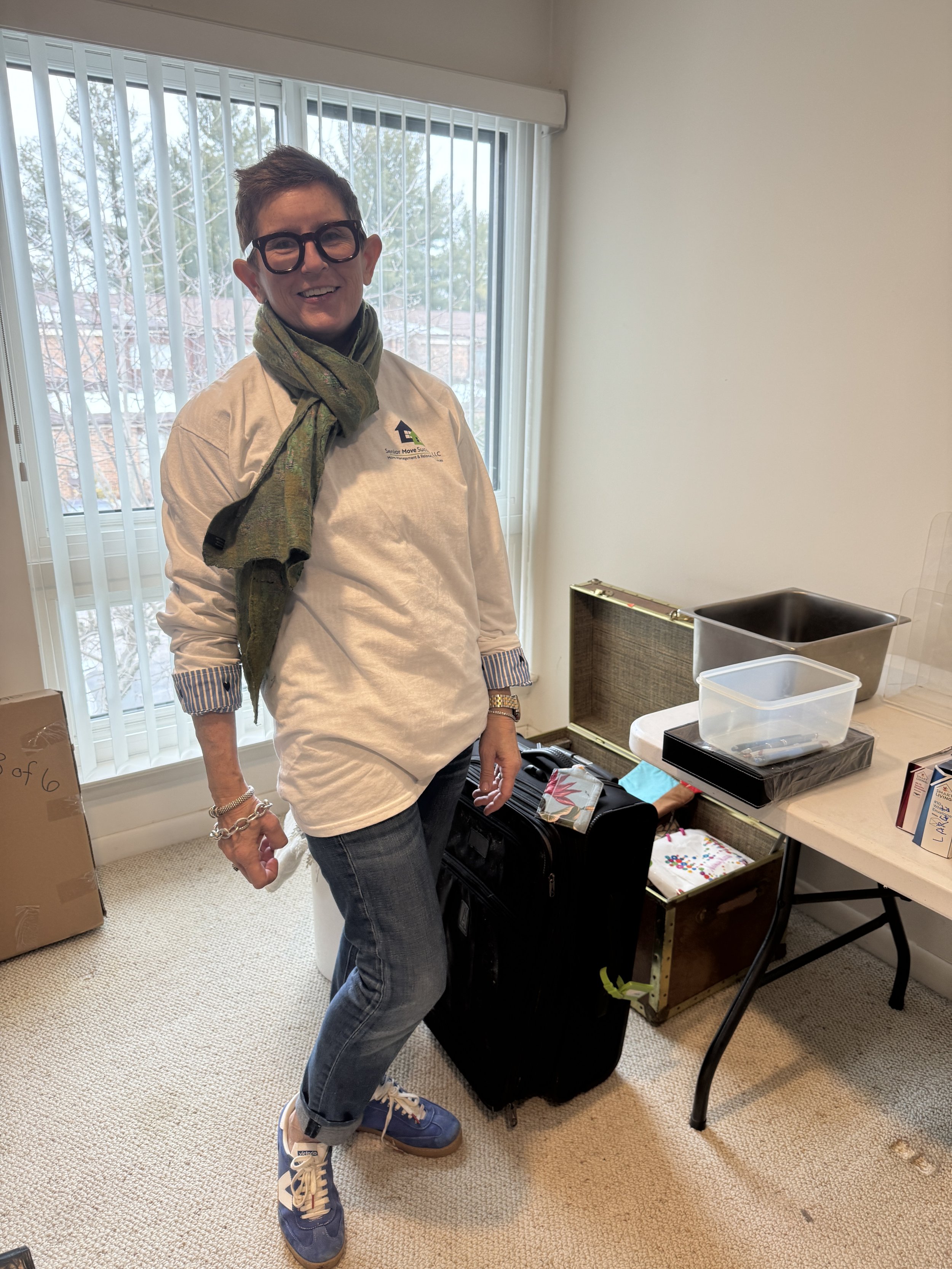 A smiling woman wearing glasses, a white long-sleeve shirt, blue jeans, and blue sneakers, standing in a room next to a table with various items and a suitcase.
