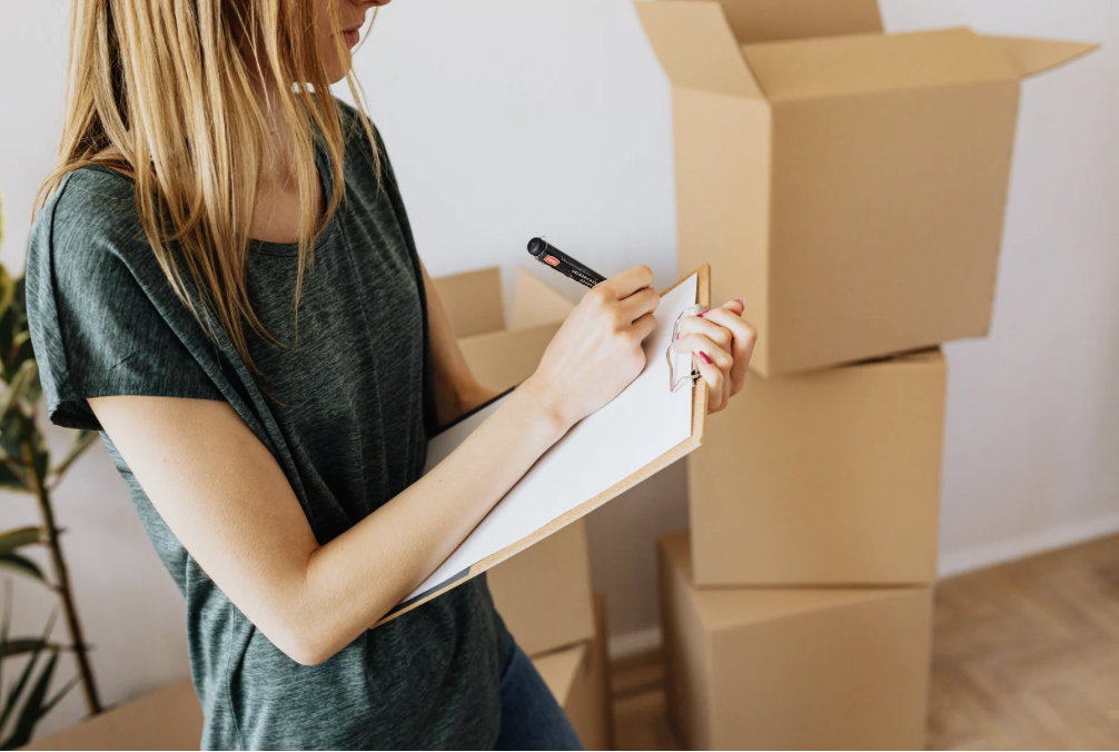 A woman writing on a clipboard with a marker, surrounded by cardboard boxes.