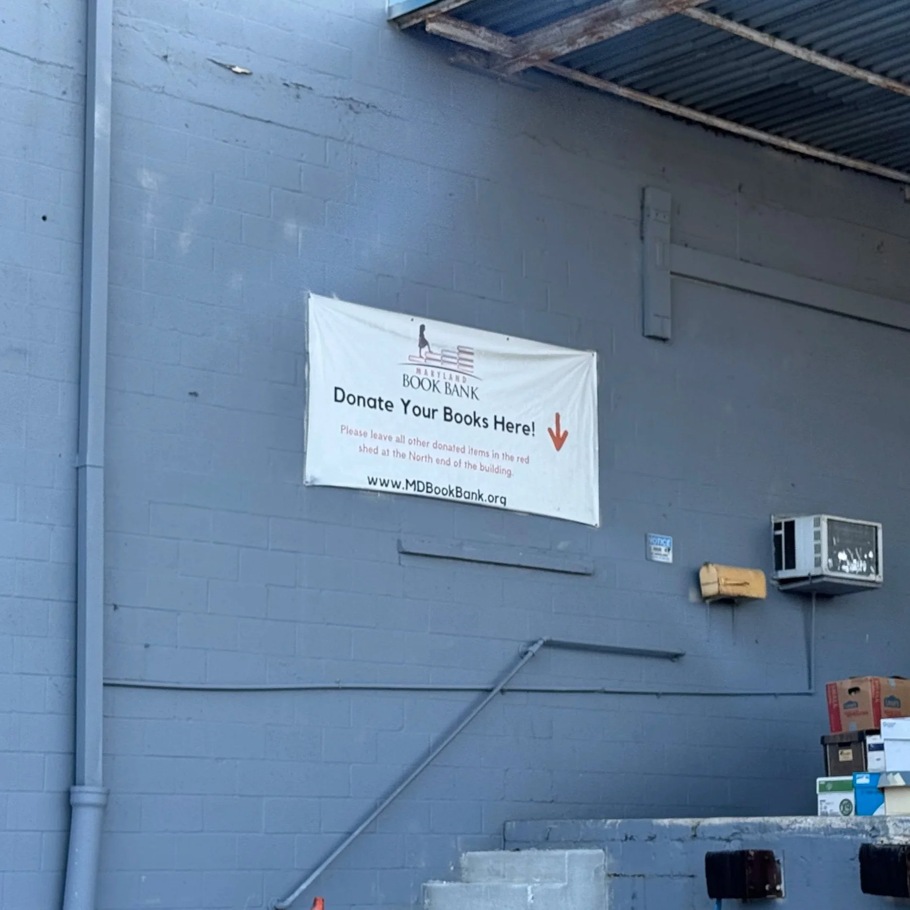 A white banner on a blue brick wall with the logo of Maryland Book Bank, calling for book donations. The banner instructs to leave books in a designated red shed at the north end of the building and provides the website www.MDBookBank.org.