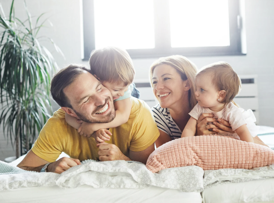A happy family of four playing and laughing on their bed in a bright, sunlit bedroom.