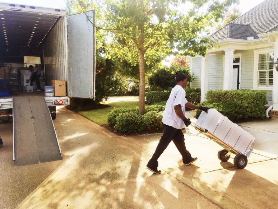 A person moving boxes on a hand truck away from a moving truck parked on a driveway. The scene is outdoors in front of a house with a well-kept yard and trees, during sunny weather.