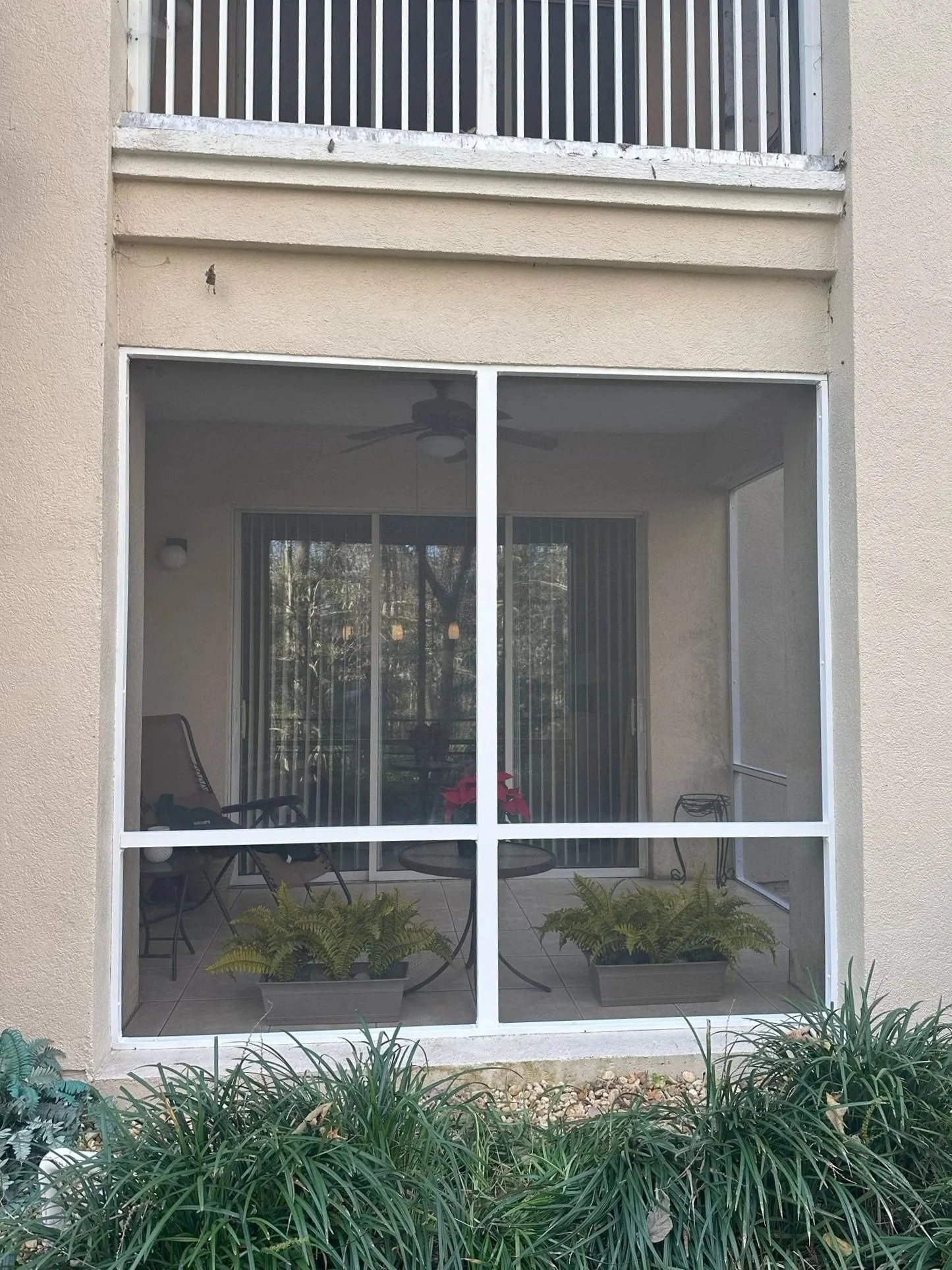 View through a screened porch with patio furniture, potted plants, and a nearby garden with green plants and rocks, on a sunny day.