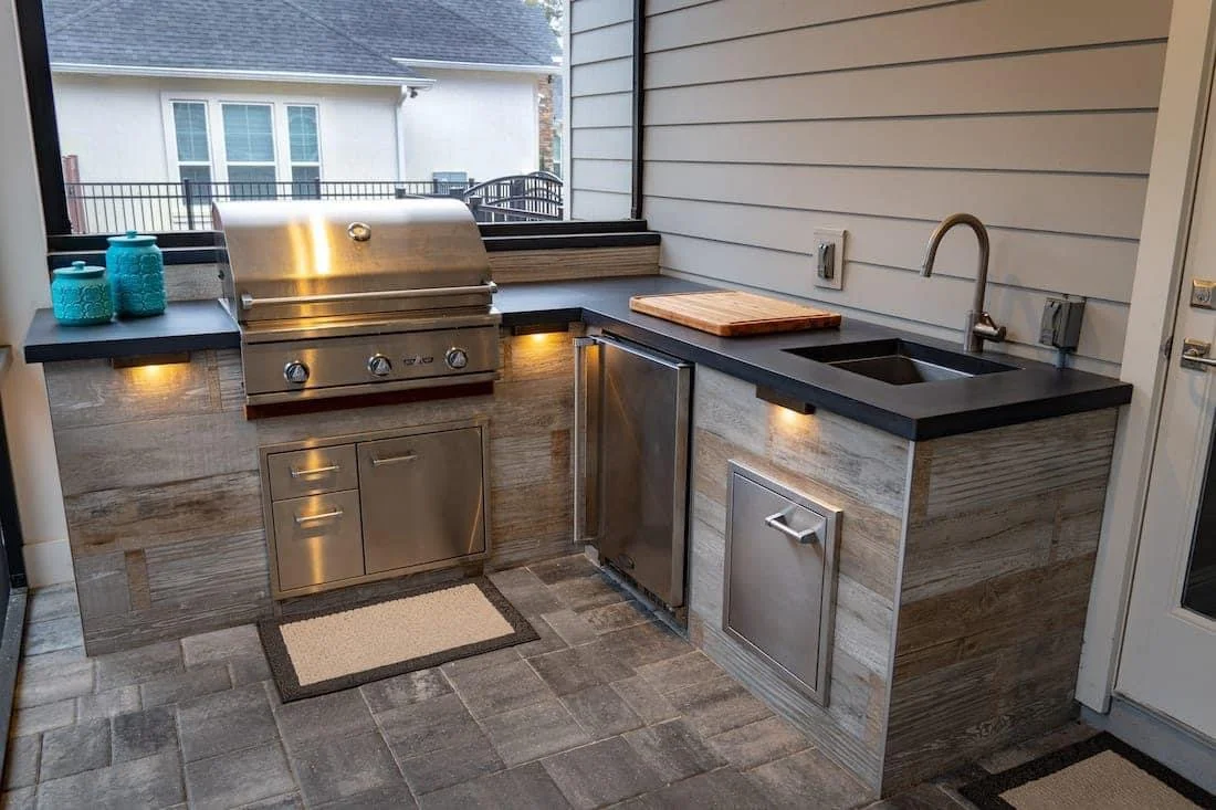 Outdoor kitchen with stainless steel grill, wooden countertops, and a black sink with a curved faucet, set against gray wooden paneling.