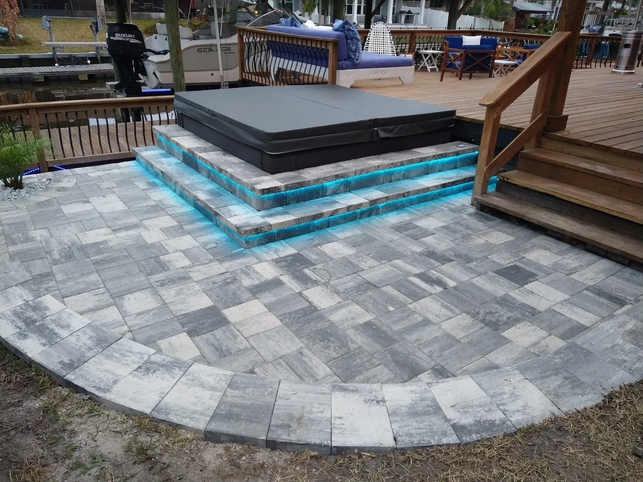 Outdoor patio area with a hot tub on a raised brick platform beside a deck with stairs. Surrounding the platform are gray paver bricks with blue LED lighting underneath the steps.