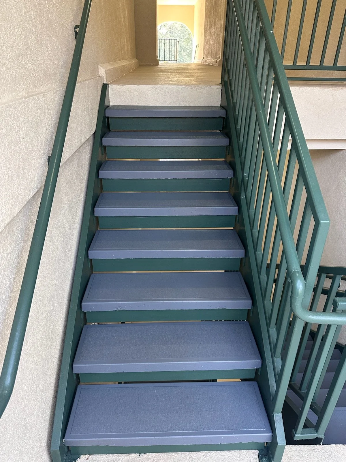 Exterior stairway with gray steps and green railings leading to a balcony at a building, with an archway and trees visible in the background.
