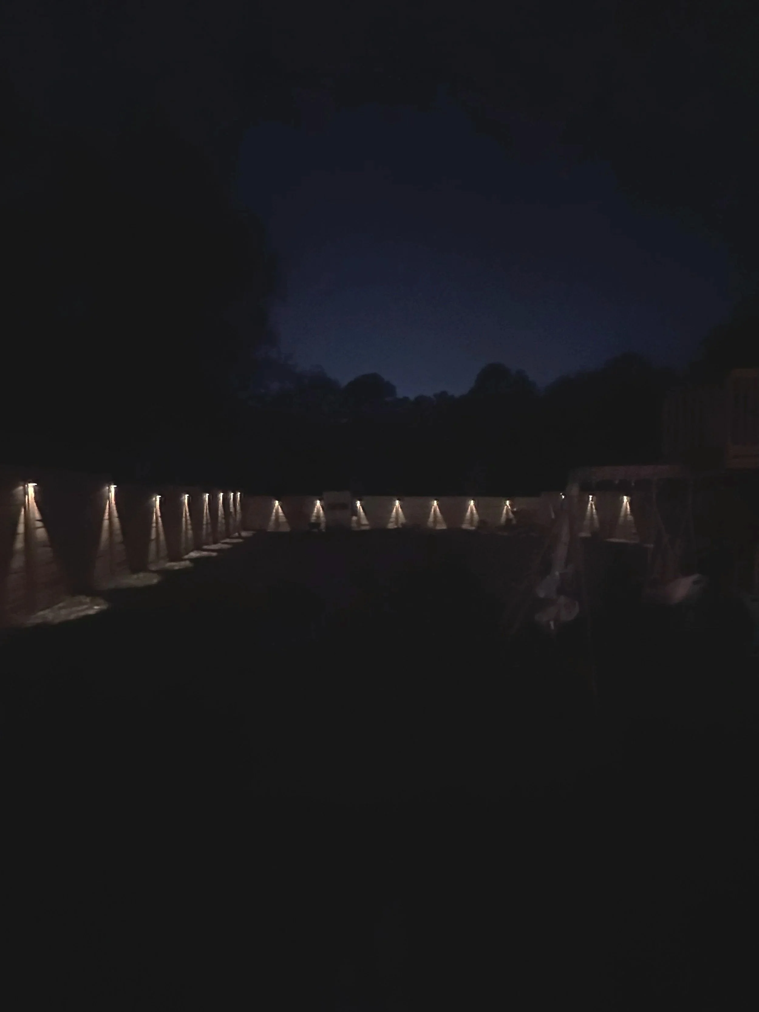 Dark night sky with clouds, distant trees, and a string of lit lamps or lights reflecting on a body of water.