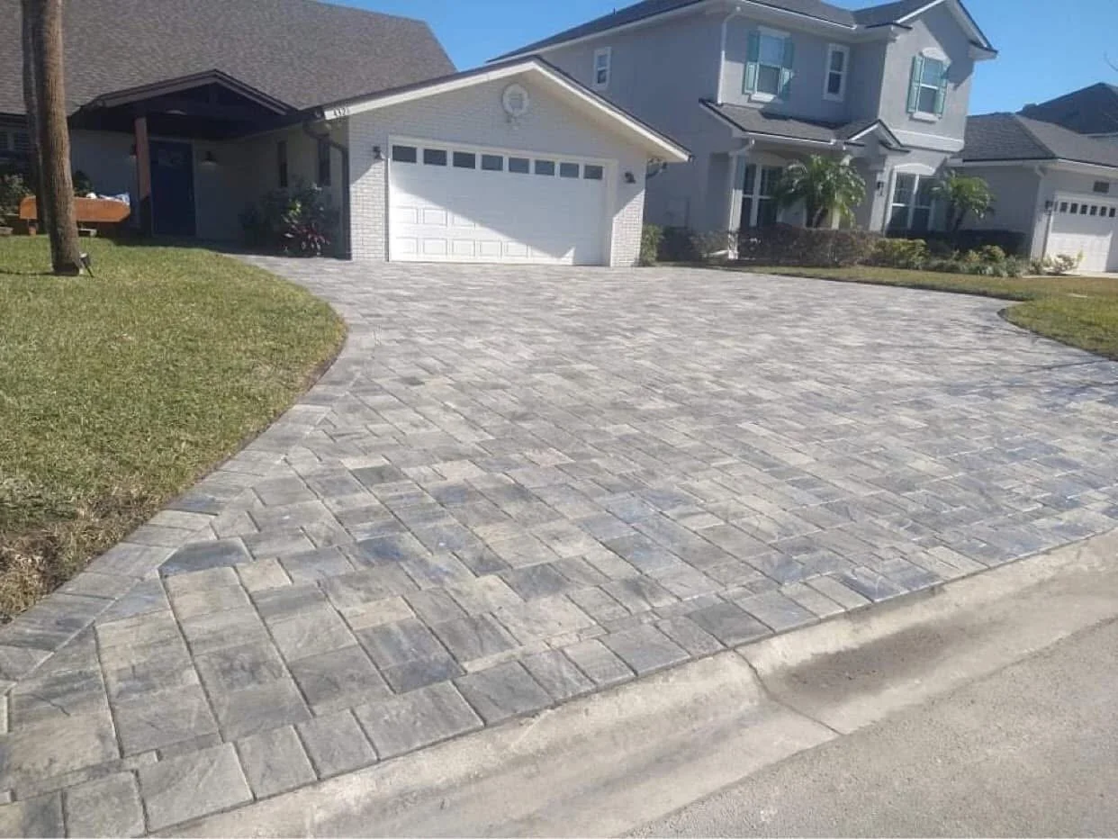 A paved driveway in front of a two-story house with a white garage door, surrounded by a lawn and a few small plants.