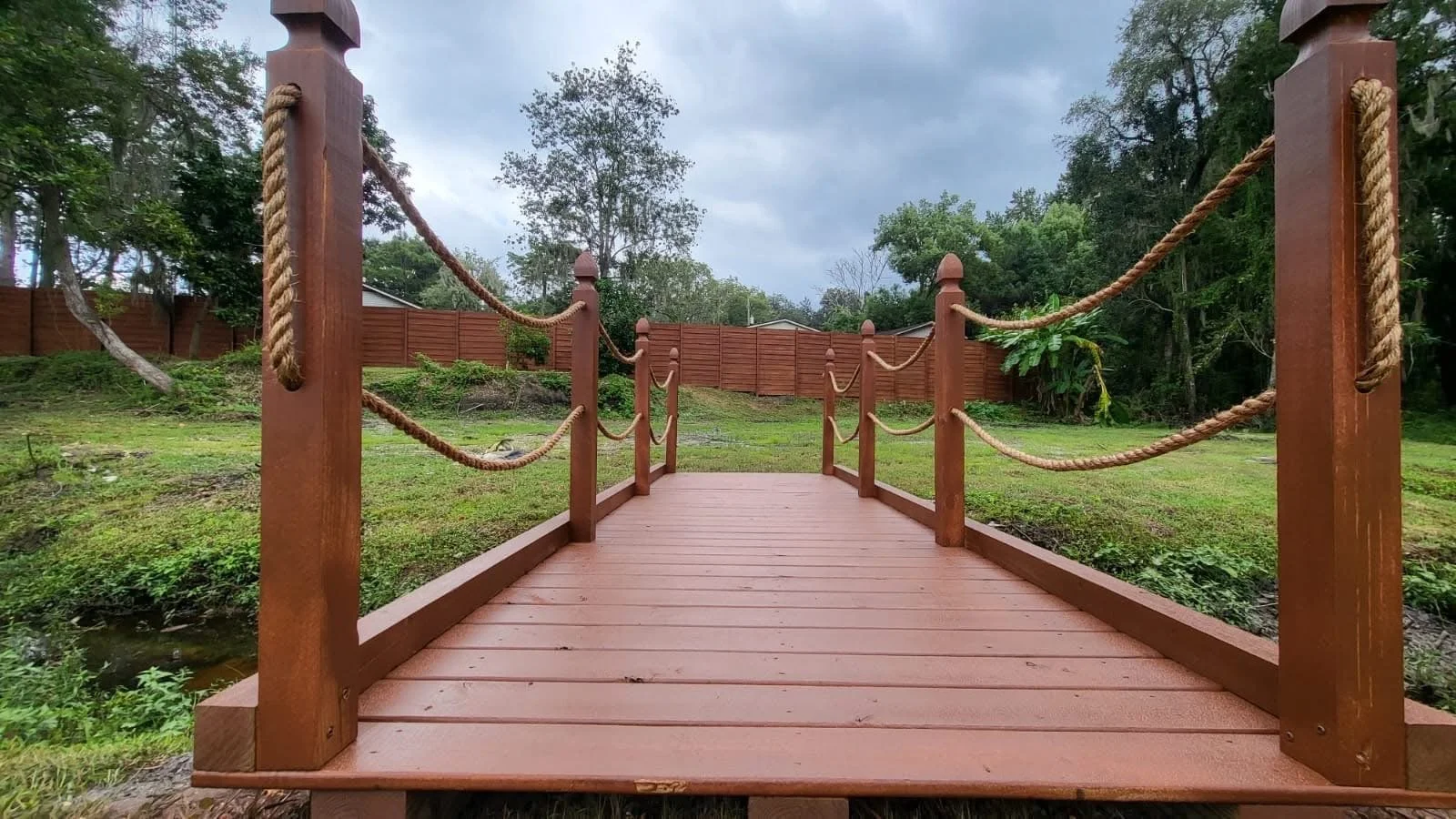 A small wooden bridge with rope railings spans over a narrow creek in a backyard with a wooden fence and trees.