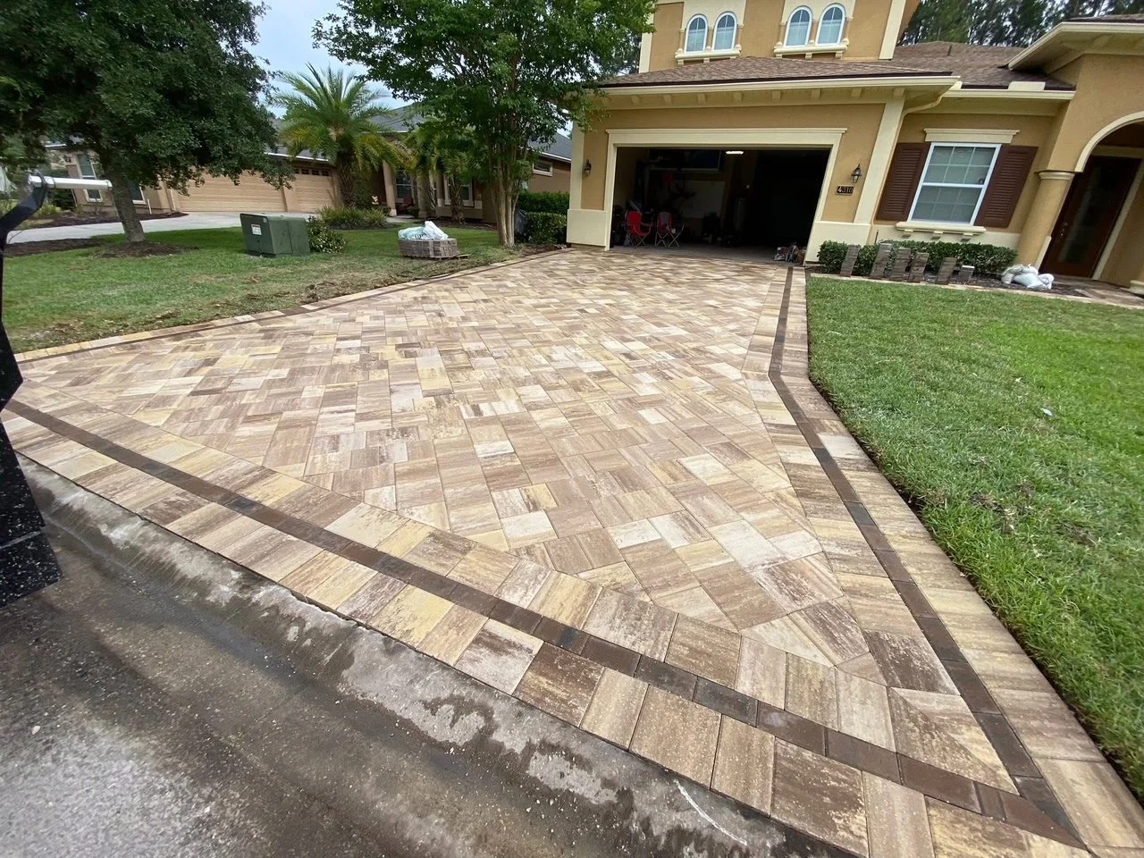 Newly paved brick driveway leading to an open garage of a beige two-story house with brown shutters, green lawn on the right, trees and neighboring houses in the background.