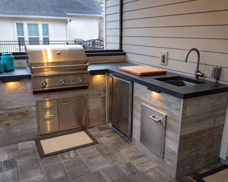 Outdoor kitchen with a stainless steel grill, a small refrigerator, a black countertop, and a sink, with a cutting board on the counter and a mat on the tiled floor.