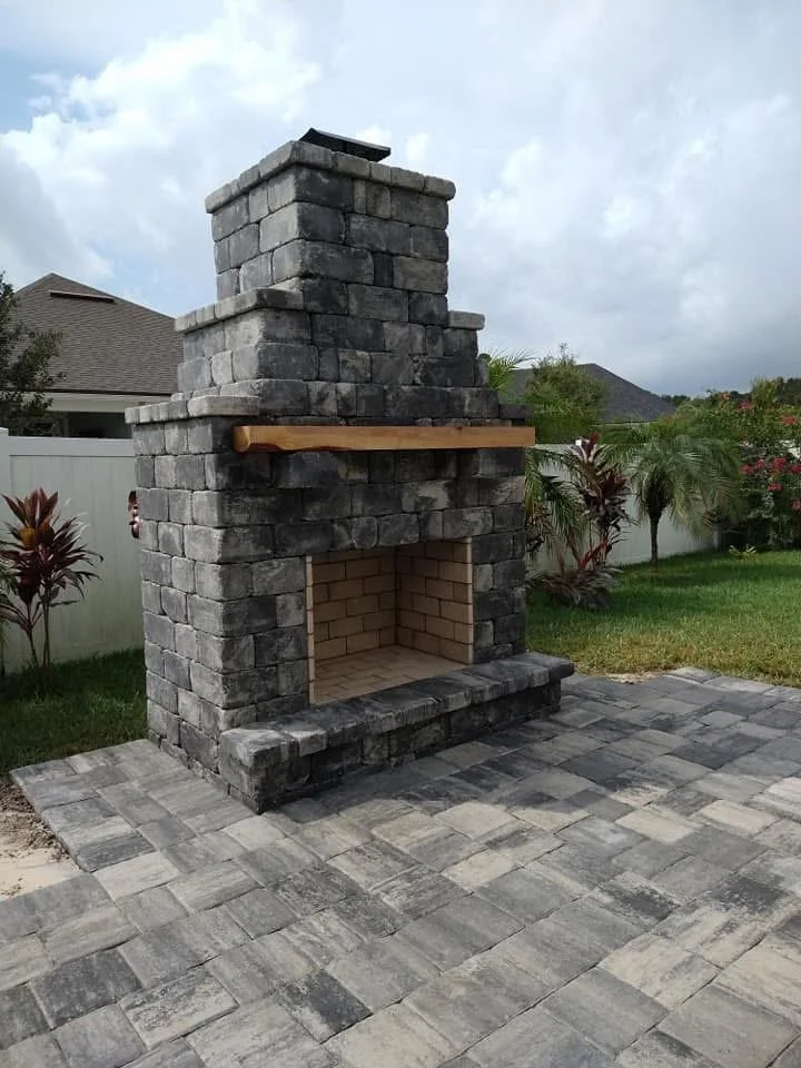 Outdoor stone fireplace with a wooden mantel, on a paved patio in a backyard with plants and a white fence.