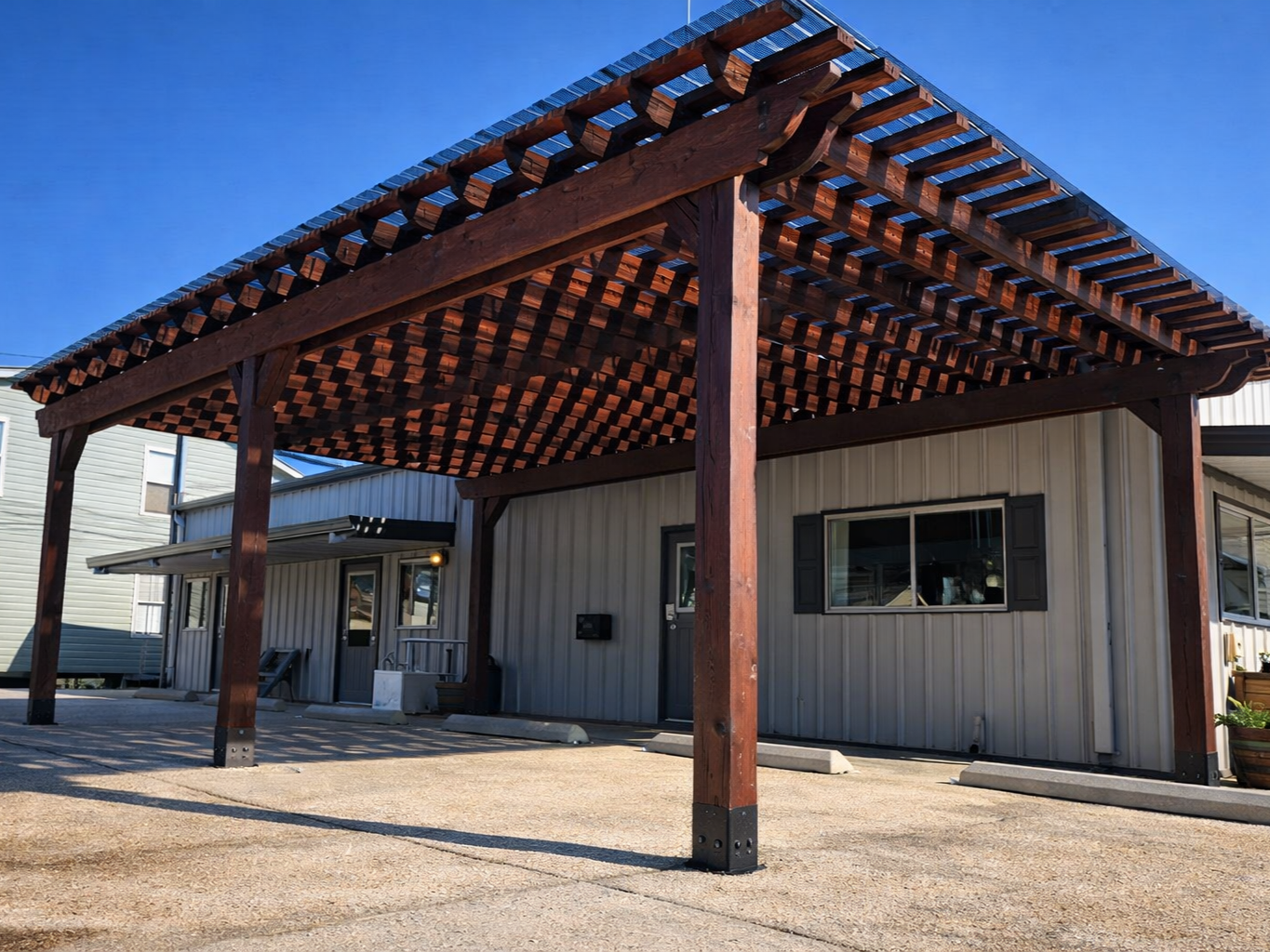 A wooden pergola attached to a small metal-clad building, with parking spaces in front and a clear blue sky overhead.