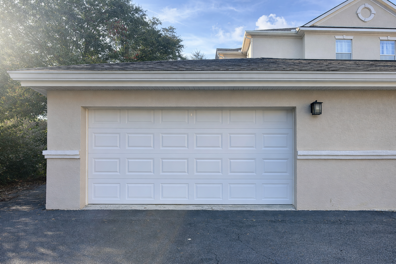 Front view of a beige suburban house with a white garage door, black wall-mounted lantern, black asphalt driveway, and part of a second story with windows and siding, with trees and blue sky in the background.