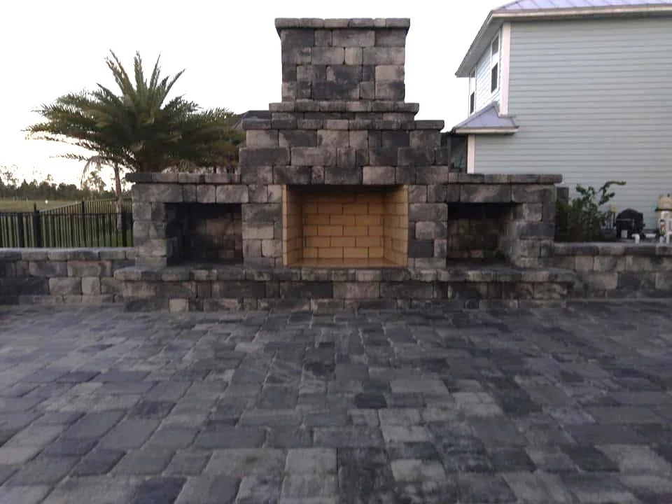 A stone outdoor fireplace with a chimney, built with large dark stones and a brick interior, situated on a stone paved patio with trees and a house in the background.