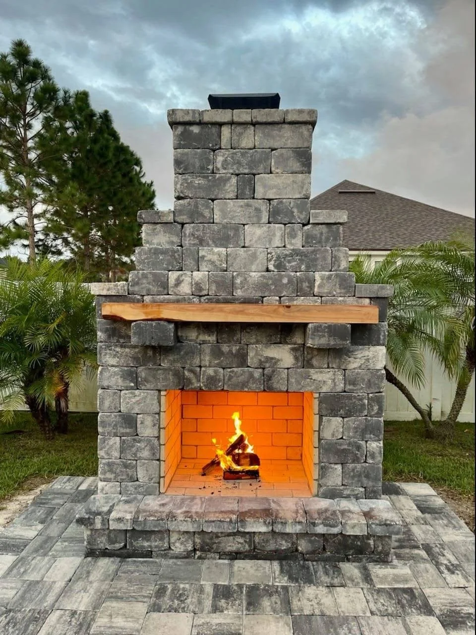 A stone outdoor fireplace with a lit fire inside, surrounded by green plants and set against a cloudy sky.