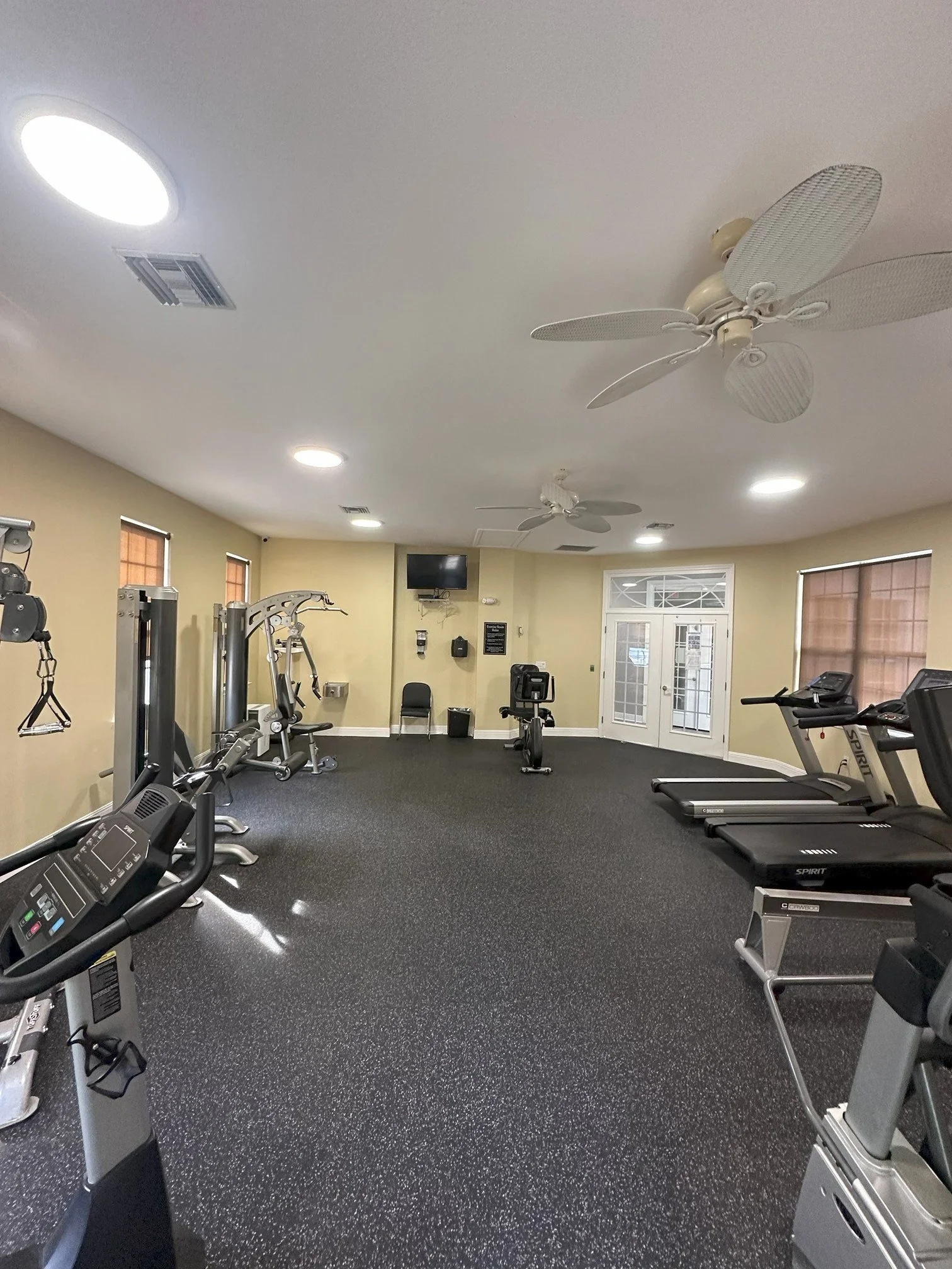 A gym with exercise machines, including treadmills and weight equipment, in a well-lit room with windows and ceiling fans.