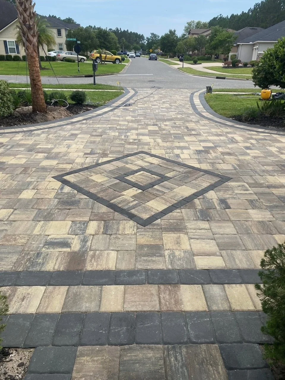 A home outdoor driveway with a decorative pattern in the pavers, leading to a residential street with houses and parked cars.