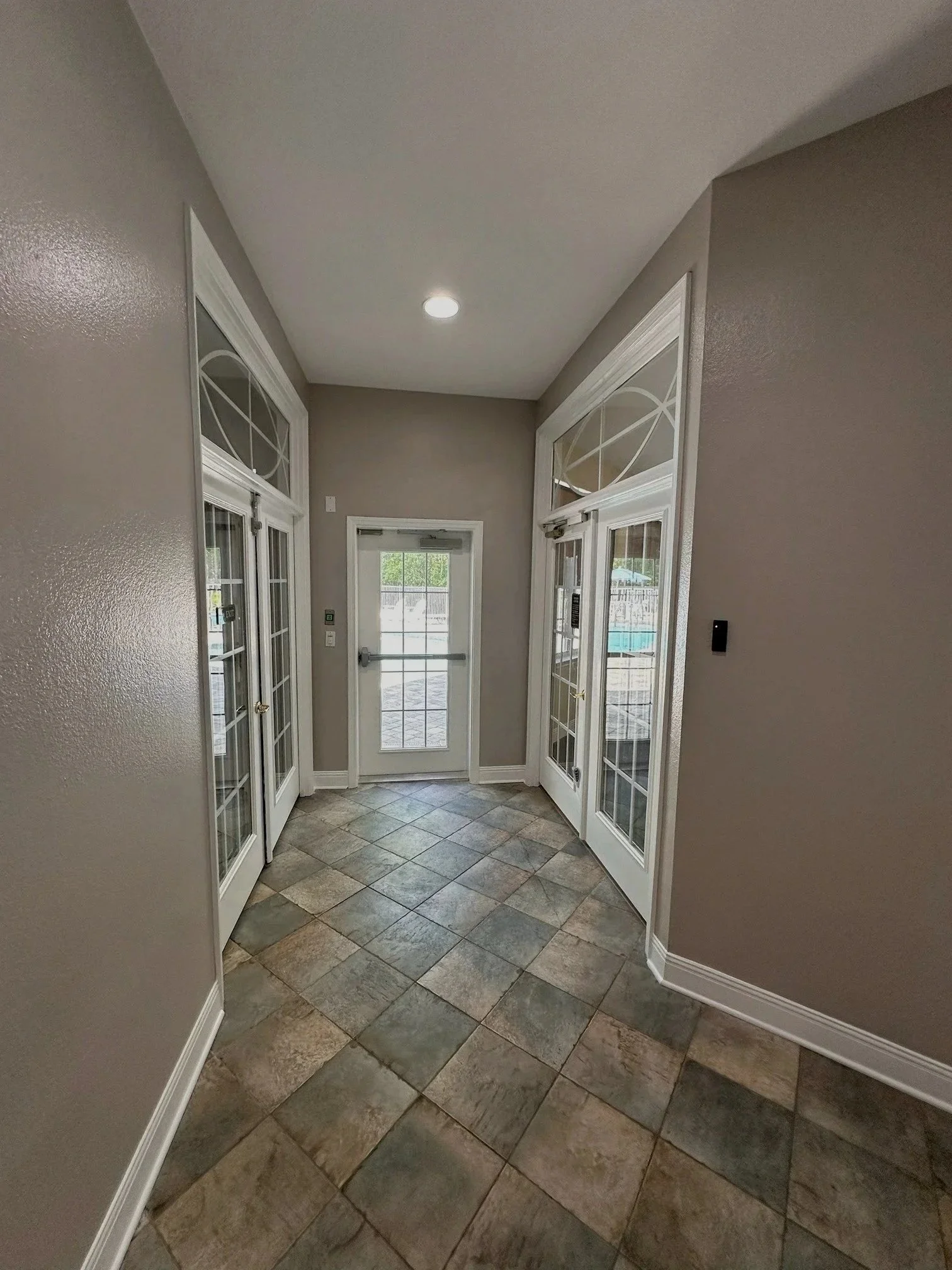 Hallway with tiled floor and beige walls, featuring glass-paneled French doors on either side and a glass door at the end, leading outside.