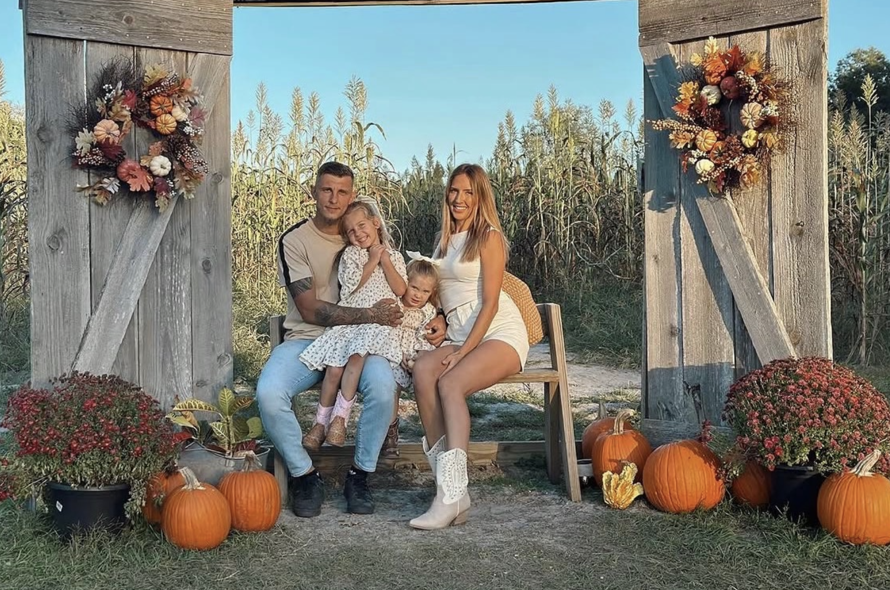 Family of four sitting on a wooden bench in front of a rustic wooden arch decorated with fall flowers and pumpkins, surrounded by outdoor fall decorations with a cornfield background under a blue sky.
