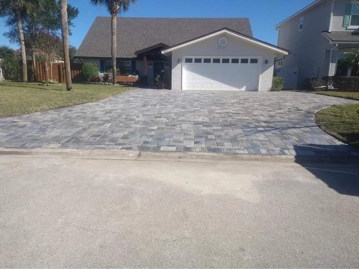 A house with a white garage door, a paved driveway, two palm trees, and a lawn with a bench on the left.