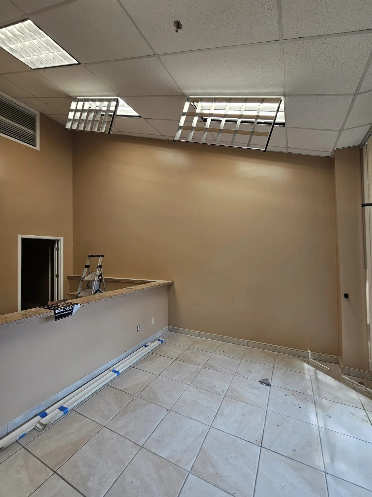 Interior room under renovation with beige walls, tiled floor, an uncovered ceiling with two ceiling tiles removed, and exposed pipes along the base of the wall.