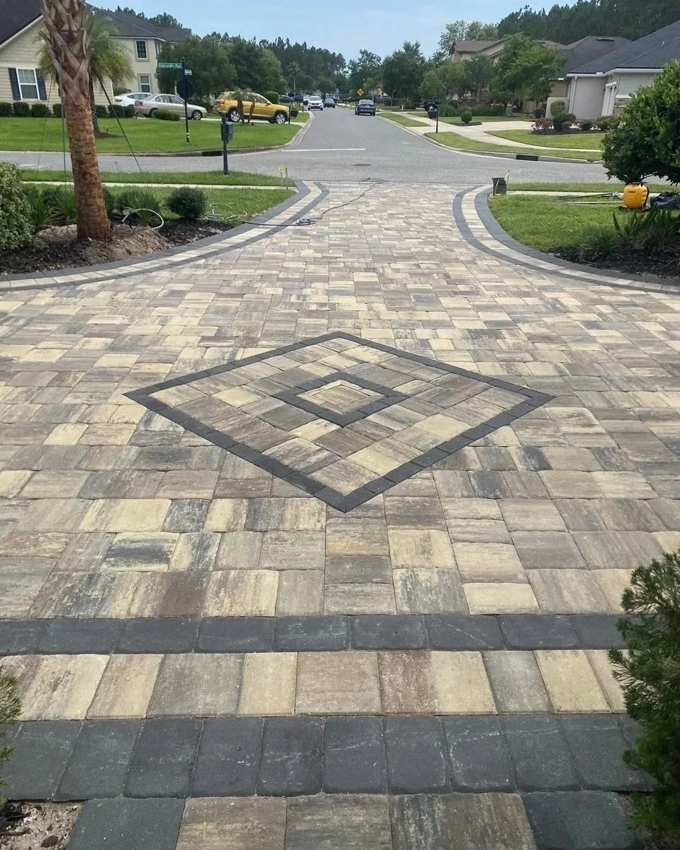 A paved driveway with a geometric pattern in the center leading to a residential street with houses, trees, and parked cars in a suburban neighborhood.