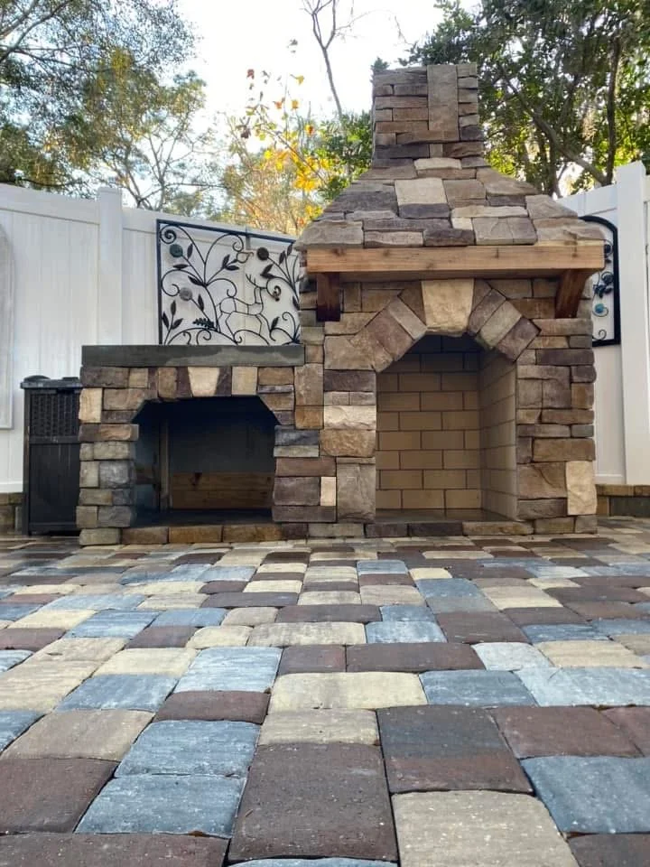 Outdoor backyard patio with a stone-built fireplace and grill area, colorful paving stones, and a white fence with decorative black metal accents, surrounded by trees.