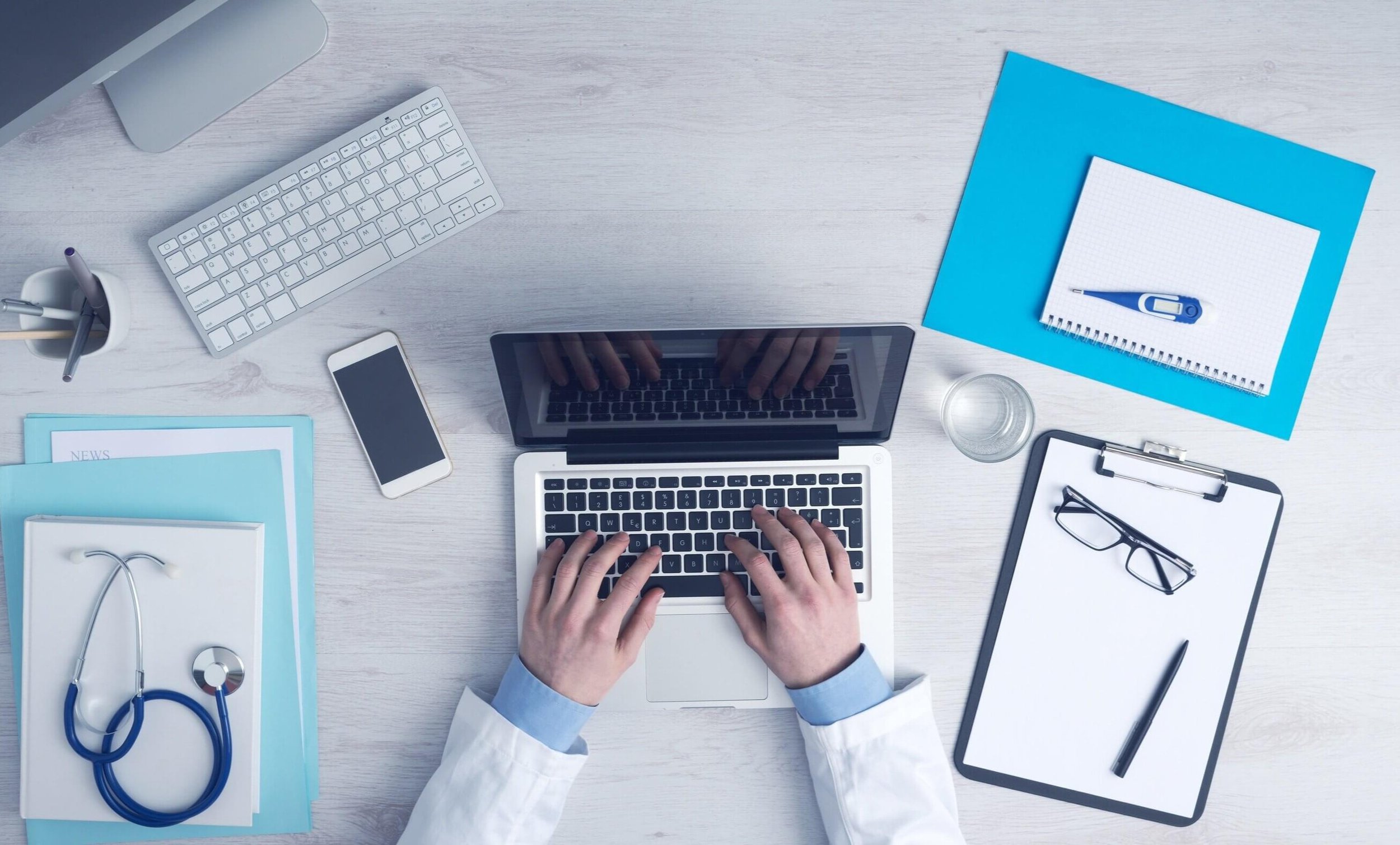 Ein Schreibtisch mit einem geöffneten Laptop, eine Person tippt auf der Tastatur, umgeben von verschiedenen Büroartikeln wie einer Tastatur, einem Smartphone, einem Wasser Glas, einem Clipboard mit einer Brille und einem Stift, sowie Ordnern und Schreibwaren.