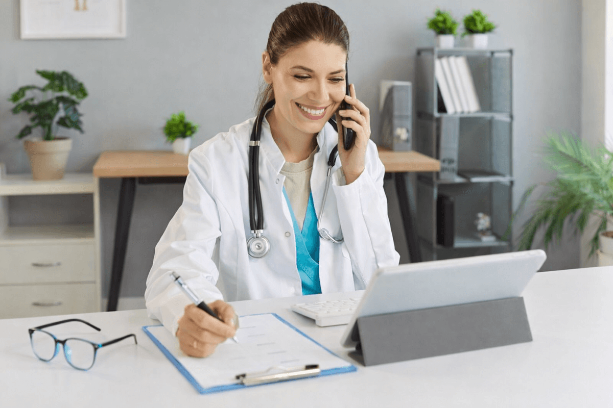 Ärztin sitzt am Schreibtisch, spricht am Telefon, schreibt auf eine medizinische Akte, vor einem Tablet, trägt ein Stethoskop, im Hintergrund Pflanzen und Ordner.