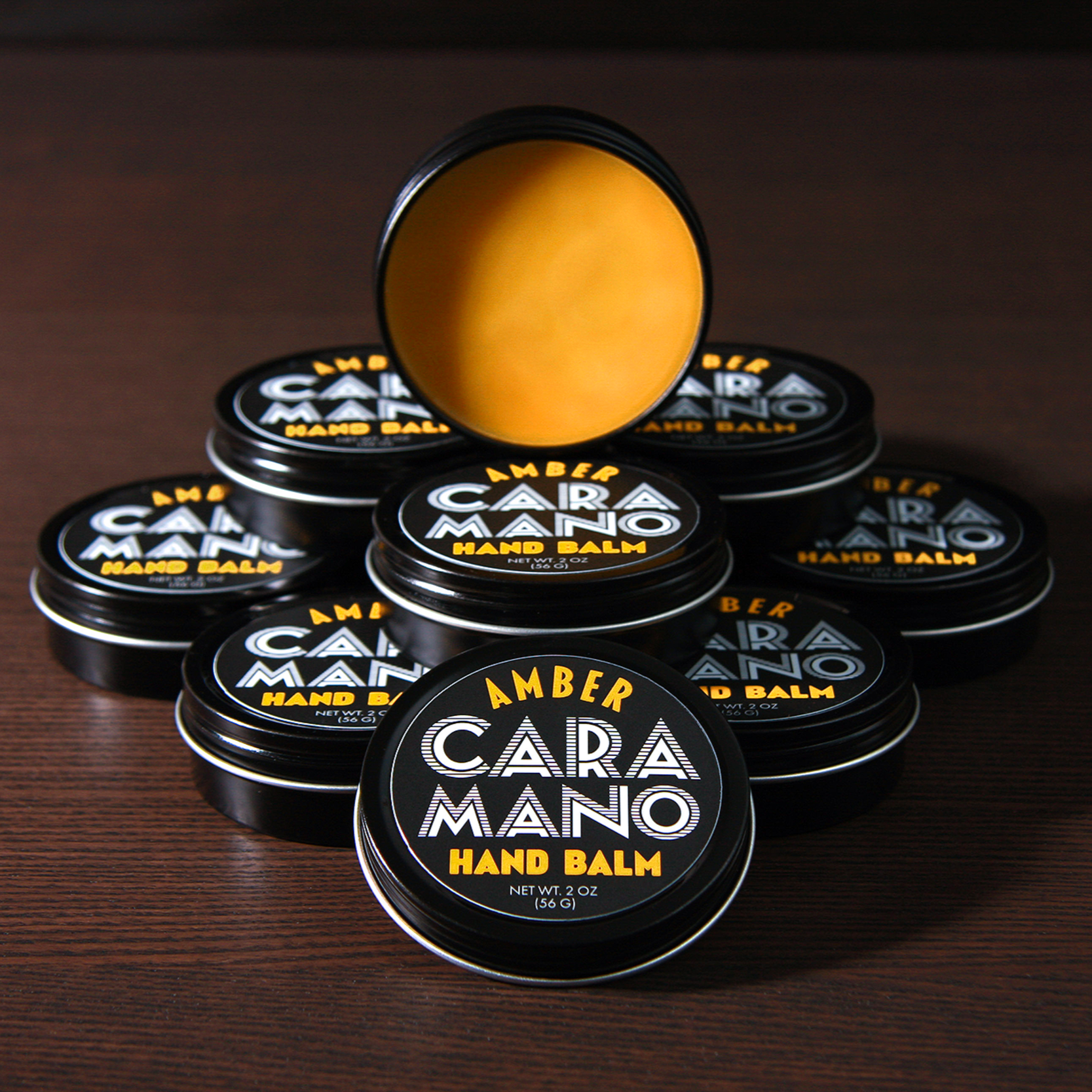 Several small black containers of Amber Caramano Hand Balm with one open container showing the yellow balm inside, arranged on a wooden surface.