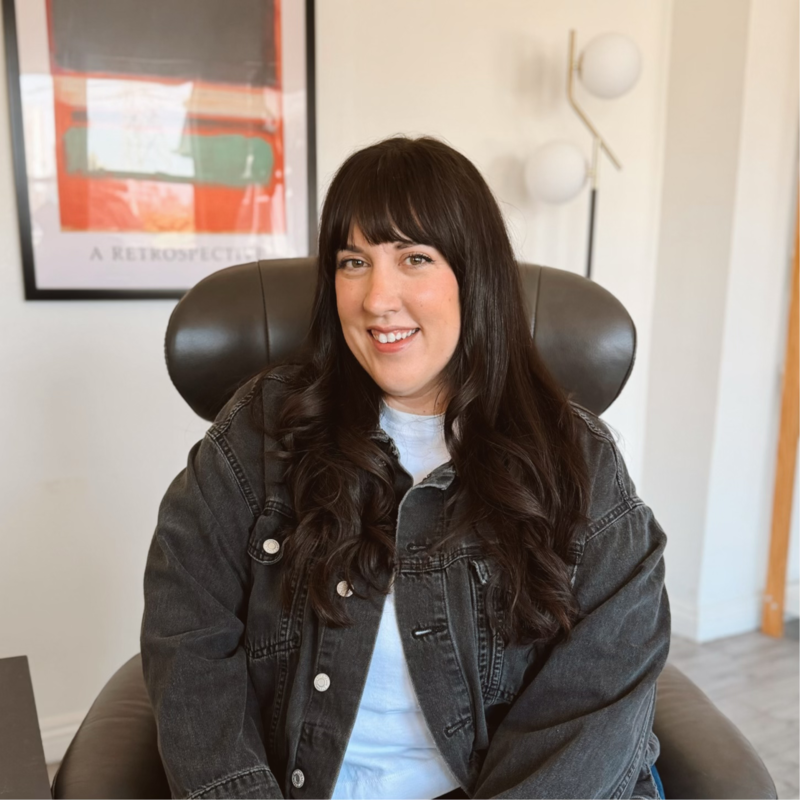 A young woman with long dark hair and bangs smiling while sitting in a gray armchair in a room with modern decor. Behind her is framed artwork and a floor lamp with two globe-shaped lights.