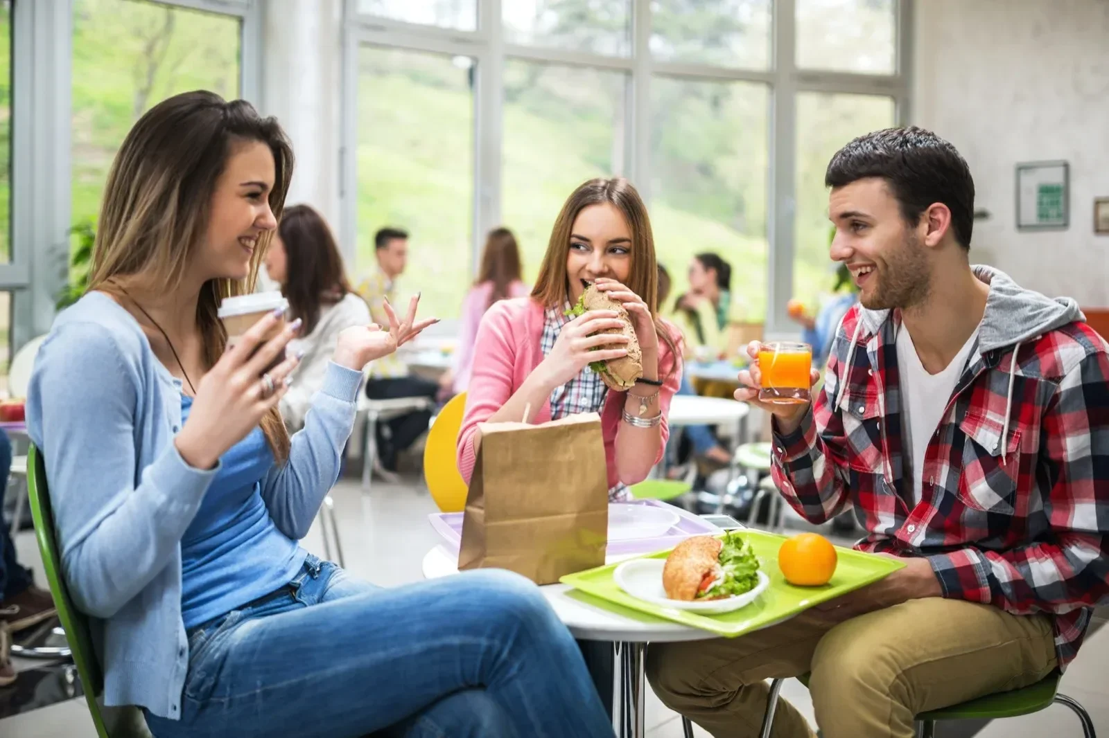 Three young people sitting together at a table in a bright cafeteria or dining area, enjoying food and drinks; two women and one man are smiling and talking, with a sandwich, salad, and orange drink on the table.